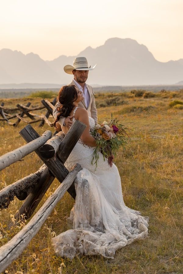 A couple in formal wedding attire embraces on a sandy beach at sunset with pink and orange skies.