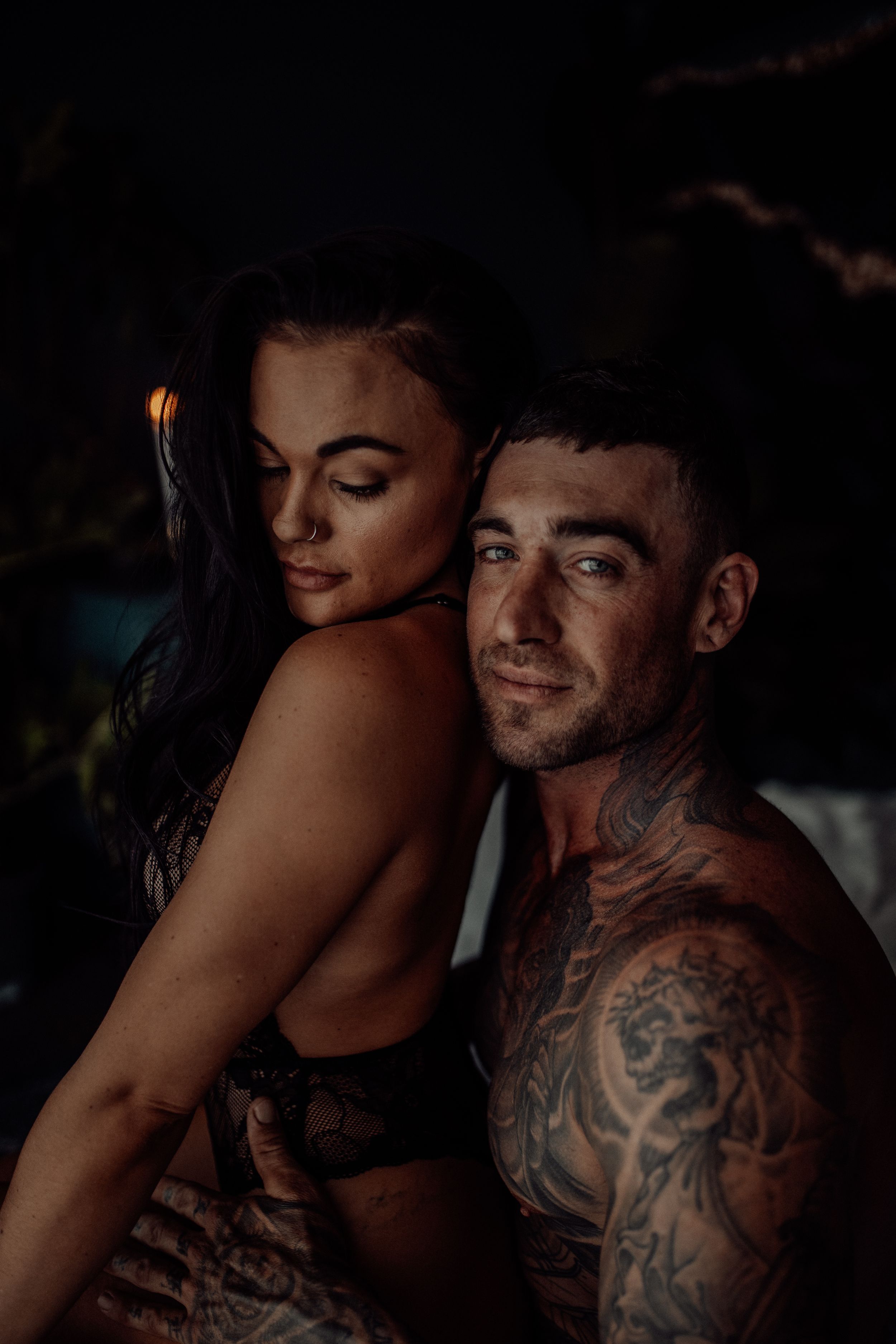 A moody, intimate portrait of a couple in low light with visible tattoos and romantic posing.