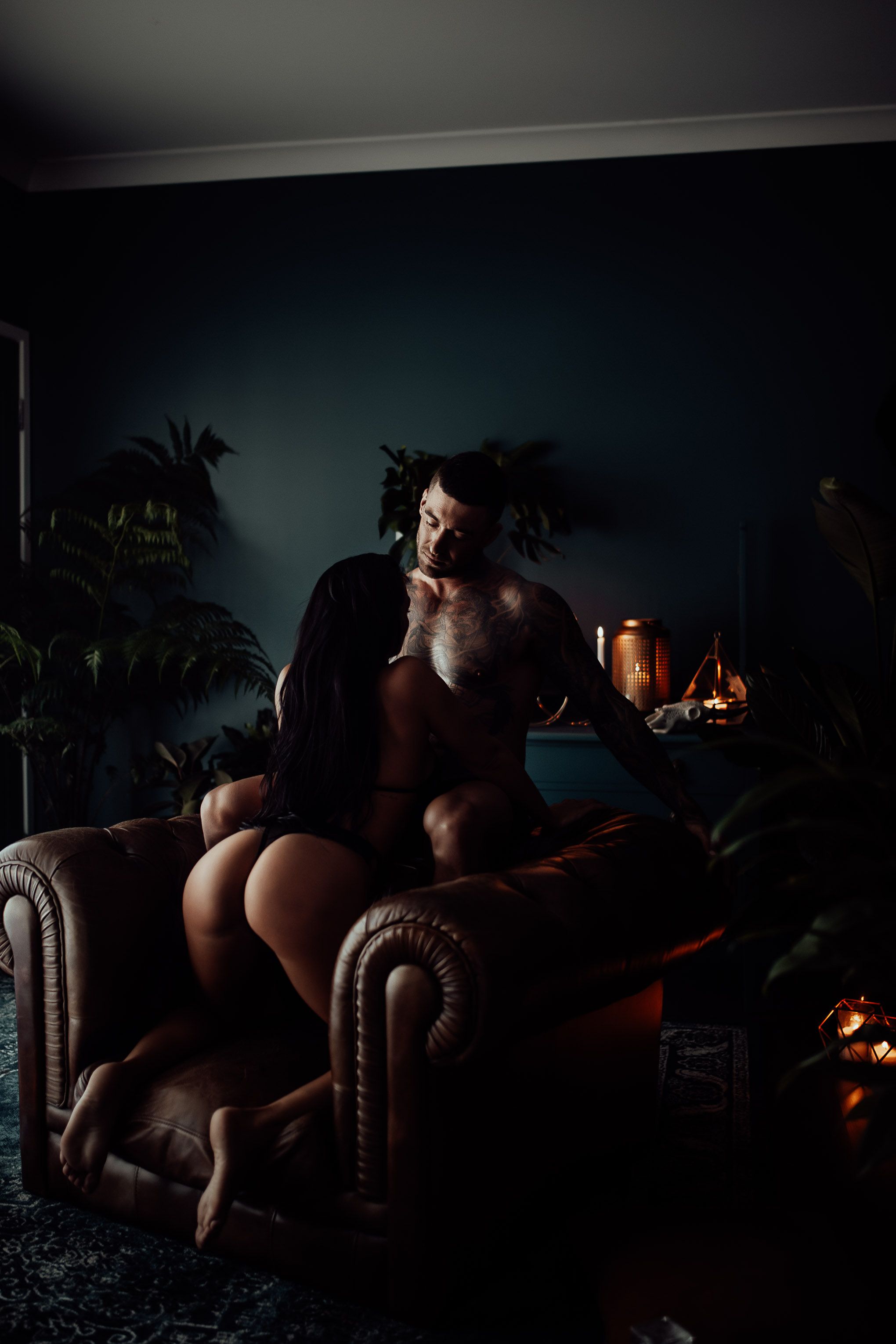 Intimate scene showing tattoo art silhouetted against dark room interior with houseplants and soft lighting.