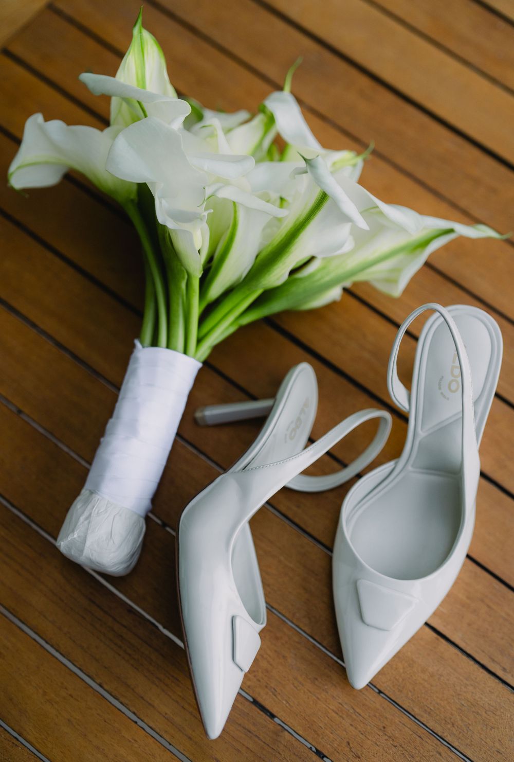 White calla lily bridal bouquet and matching white slingback heels arranged on wooden floor.