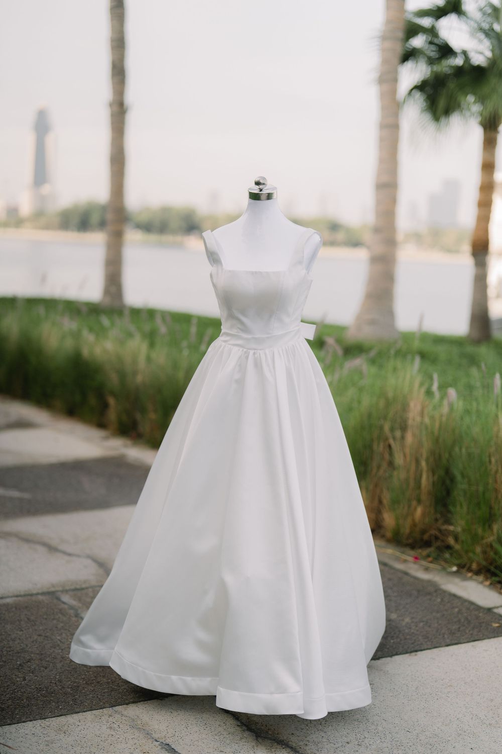 Elegant white wedding dress with square neckline and full skirt displayed on mannequin outdoors with palm trees in background.