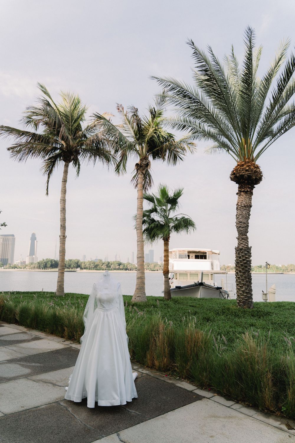 A white wedding dress stands empty against a scenic backdrop of palm trees and cityscape along a waterfront.