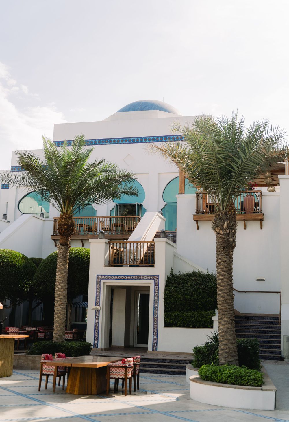 Mediterranean-style white building with blue dome and palm trees framing the entrance courtyard.