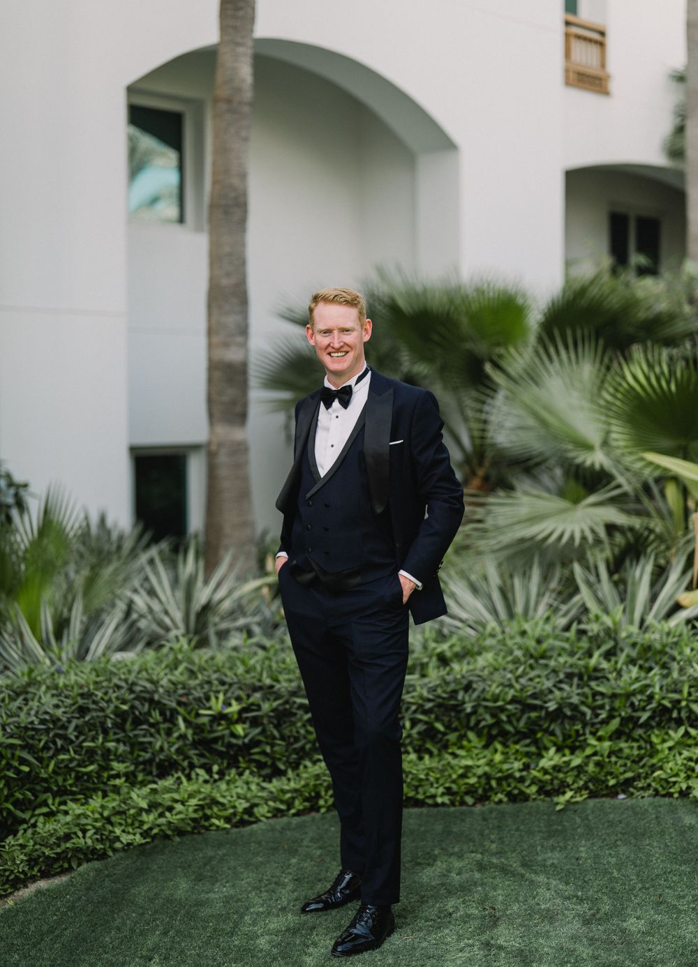 A smiling person in a black tuxedo and bowtie stands in front of a white Mediterranean-style building with palm plants.