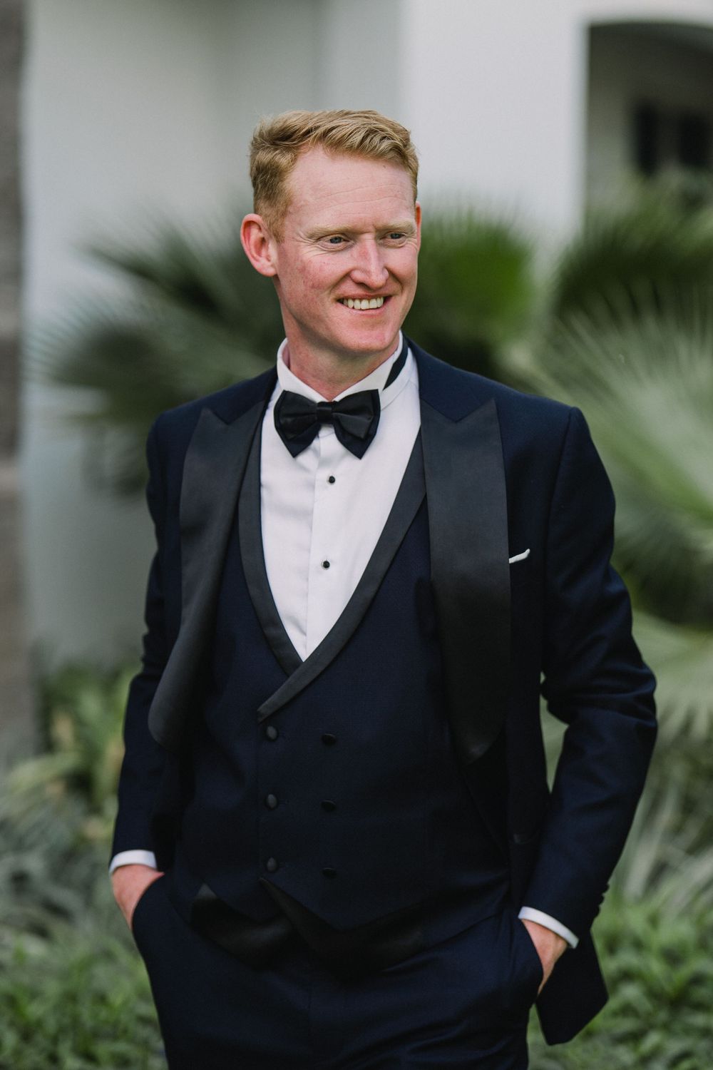 A person in a classic black tuxedo with bow tie smiles while standing near tropical foliage.