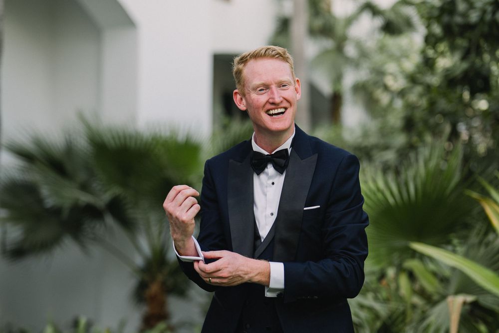 Groom in classic black tuxedo and bowtie laughs joyfully while adjusting cufflinks in outdoor garden setting.