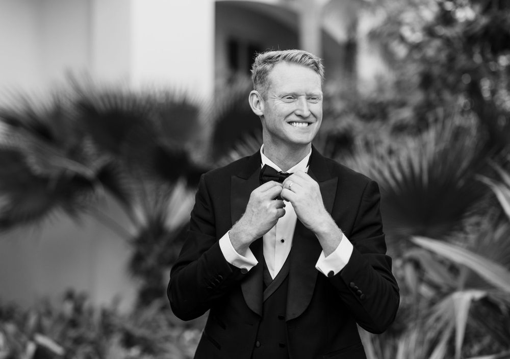 A black and white photograph shows a well-dressed man in a tuxedo adjusting his bow tie while smiling outdoors.