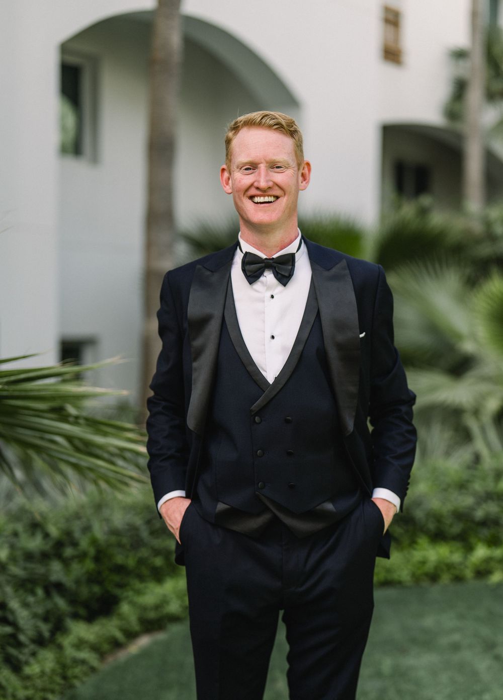 A person in a classic black tuxedo with bowtie poses outdoors in front of white arched architecture and tropical plants.