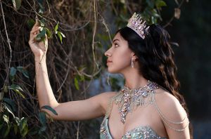 A woman in a sparkling gown and crystal crown reaches for leaves among forest vines in an enchanting fairy tale scene.