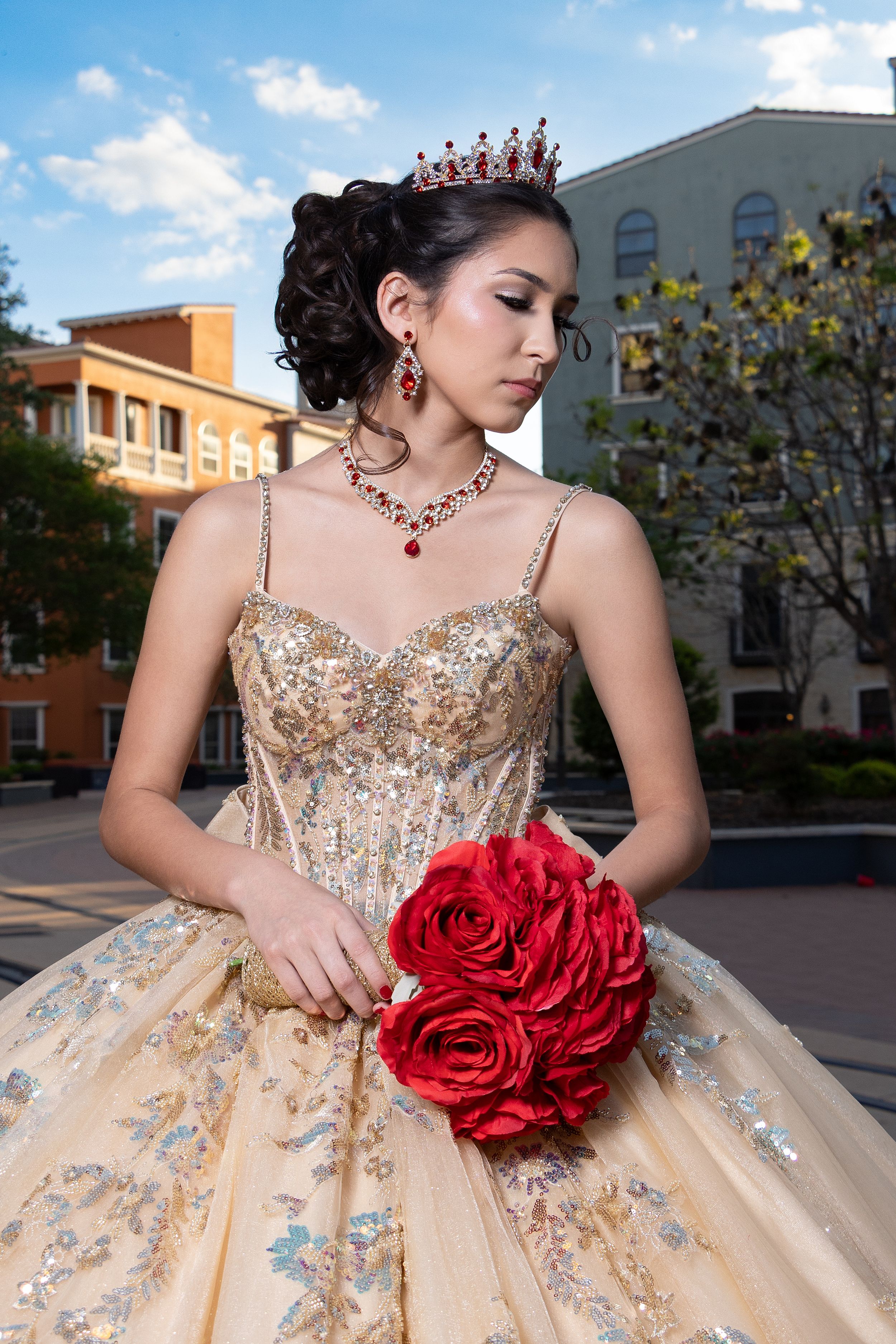 Young woman in gold quinceañera gown with red rose bouquet, ruby crown and jewelry, posing outdoors.