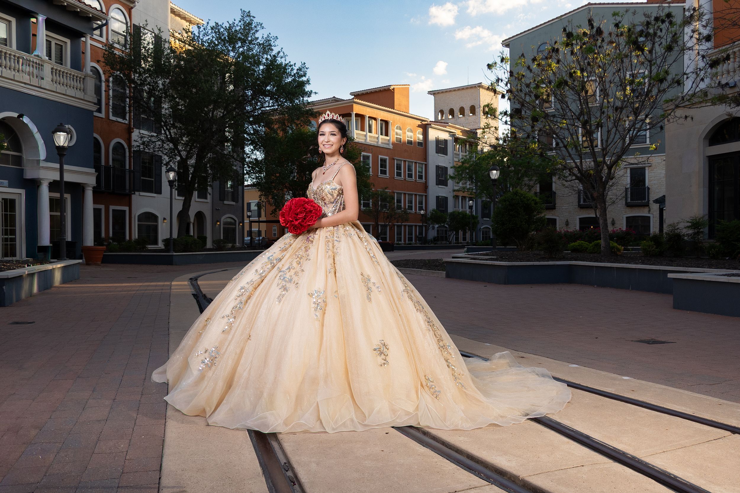 Young woman in a golden ball gown quinceañera dress holding red roses in an urban plaza setting.