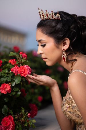 Young woman wearing a gold crown and earrings smells red roses in a garden, dressed in an elegant gold gown.