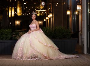 Young woman in a yellow ball gown with pink floral details and tiara poses outdoors at night under warm string lights.