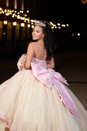 Young woman in a sparkly ballgown with pink bow and tiara smiles over her shoulder at a night event.