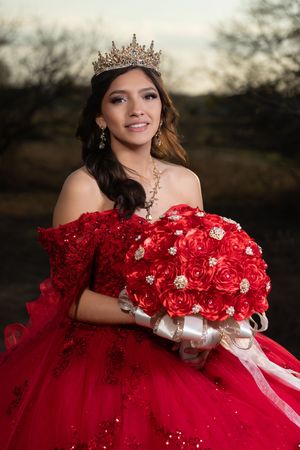 Young Latina in red quinceañera gown, gold crown, and matching rose bouquet poses outdoors at golden hour.