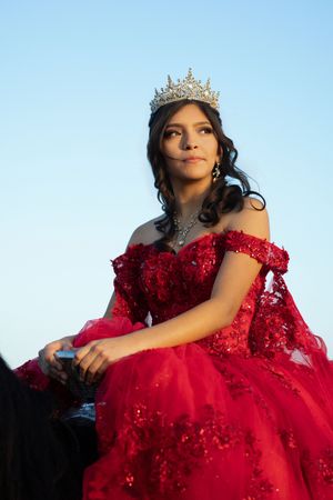Young woman in red quinceañera gown with crown sits elegantly outdoors against a clear blue sky.
