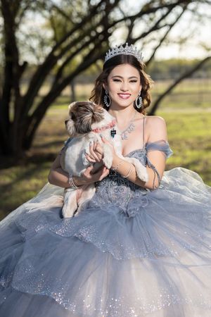 Young woman in blue ball gown and tiara holds small dog outdoors, smiling elegantly in a princess-style quinceañera dress.