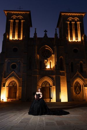 Woman in black ball gown poses before illuminated historic stone cathedral at night.