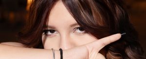 Young woman with brown hair peering over her hand, wearing stacked bracelets, with a mysterious gaze.