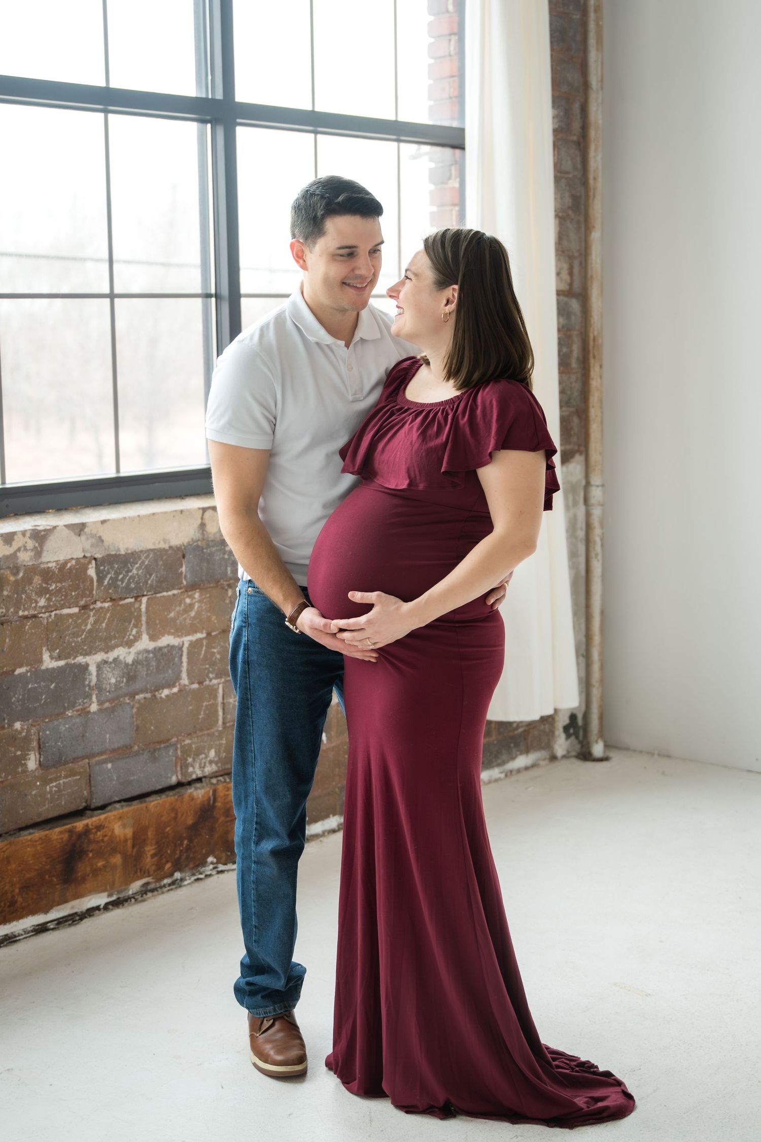 Smiling pregnant couple in studio, man in white shirt and jeans, woman in burgundy maternity gown, posing by brick wall window.