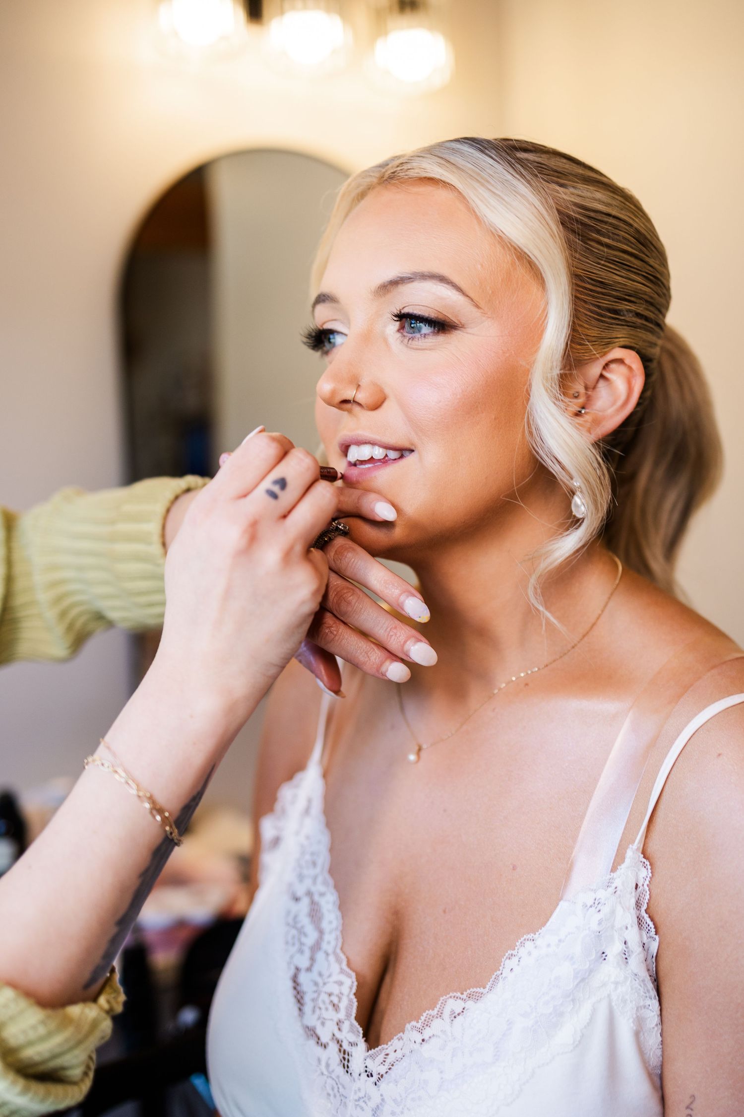Blonde bride getting earrings put on while wearing white lace dress during wedding day preparations.