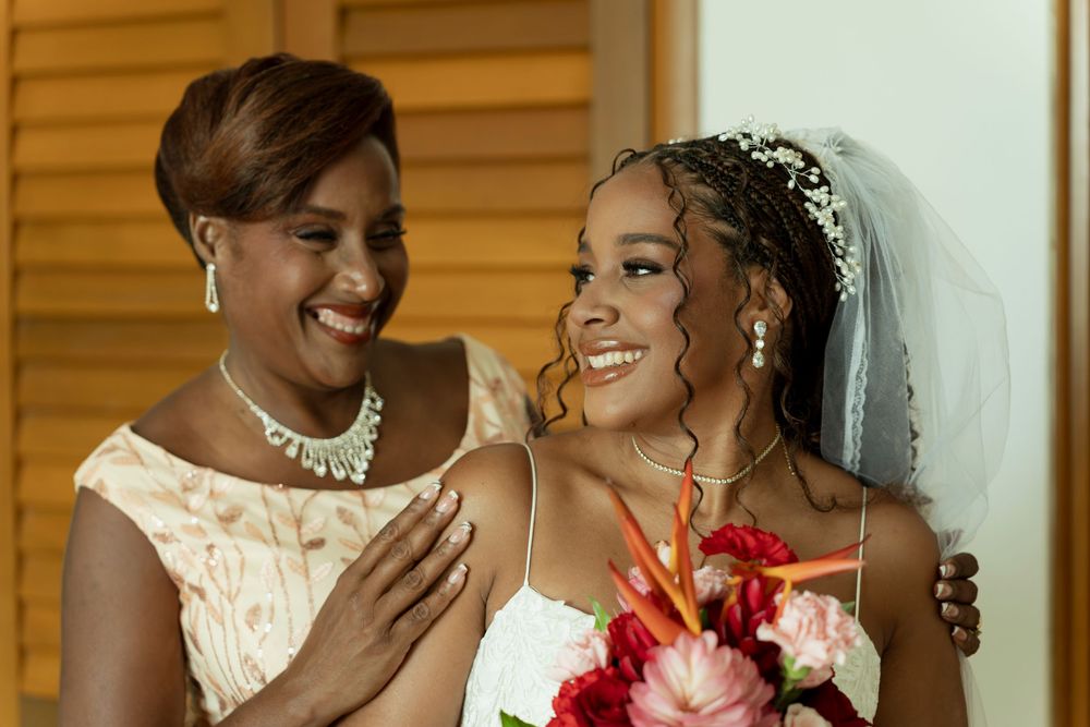 A smiling bride in a white gown and veil holds a bouquet beside an elegant woman in a lace dress at a wedding.