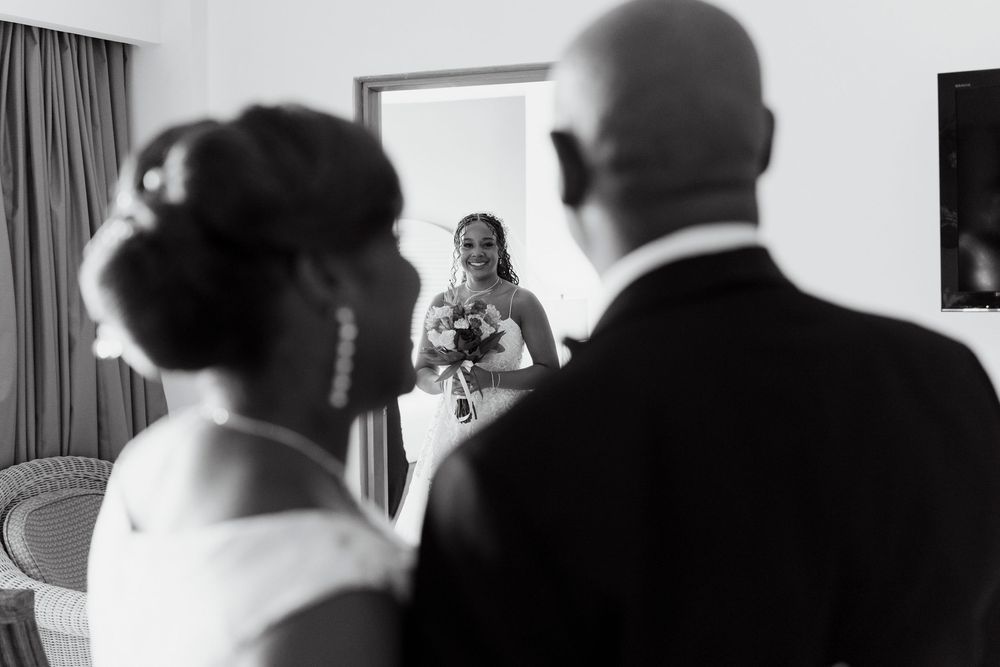 Black and white photo of a smiling bride holding bouquet, seen from behind two wedding guests in formal attire.