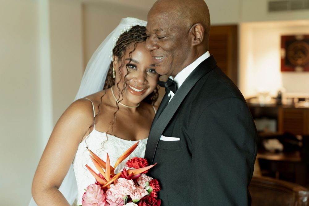 A smiling bride in a white gown and veil holds a tropical bouquet while embracing an older man in a black tuxedo.