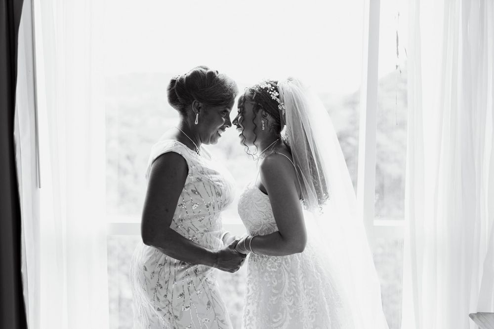Black and white photo of two women in wedding dresses holding hands by a bright window.