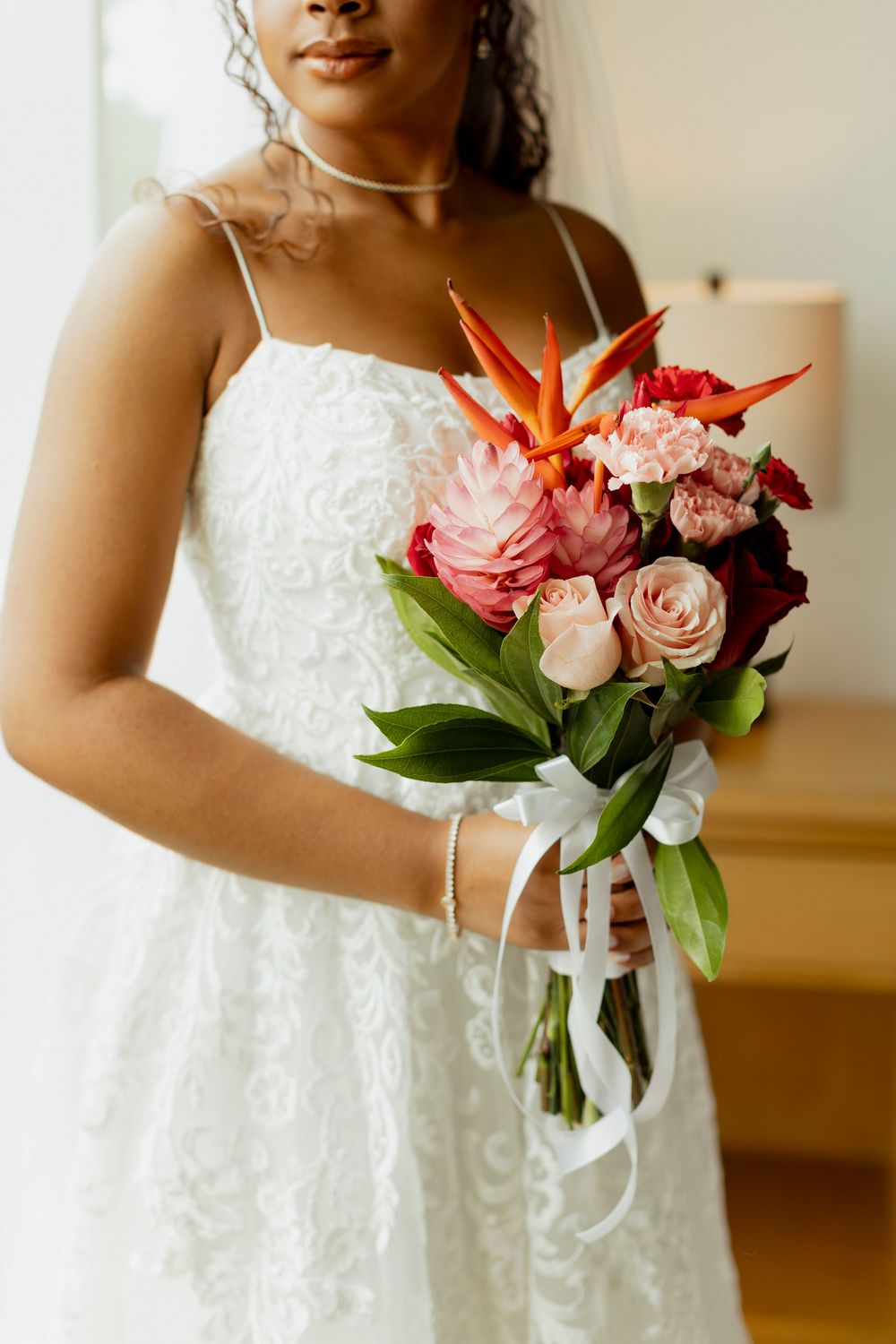 Black bride in white lace gown holds tropical wedding bouquet with birds of paradise and pink roses tied with white ribbon.
