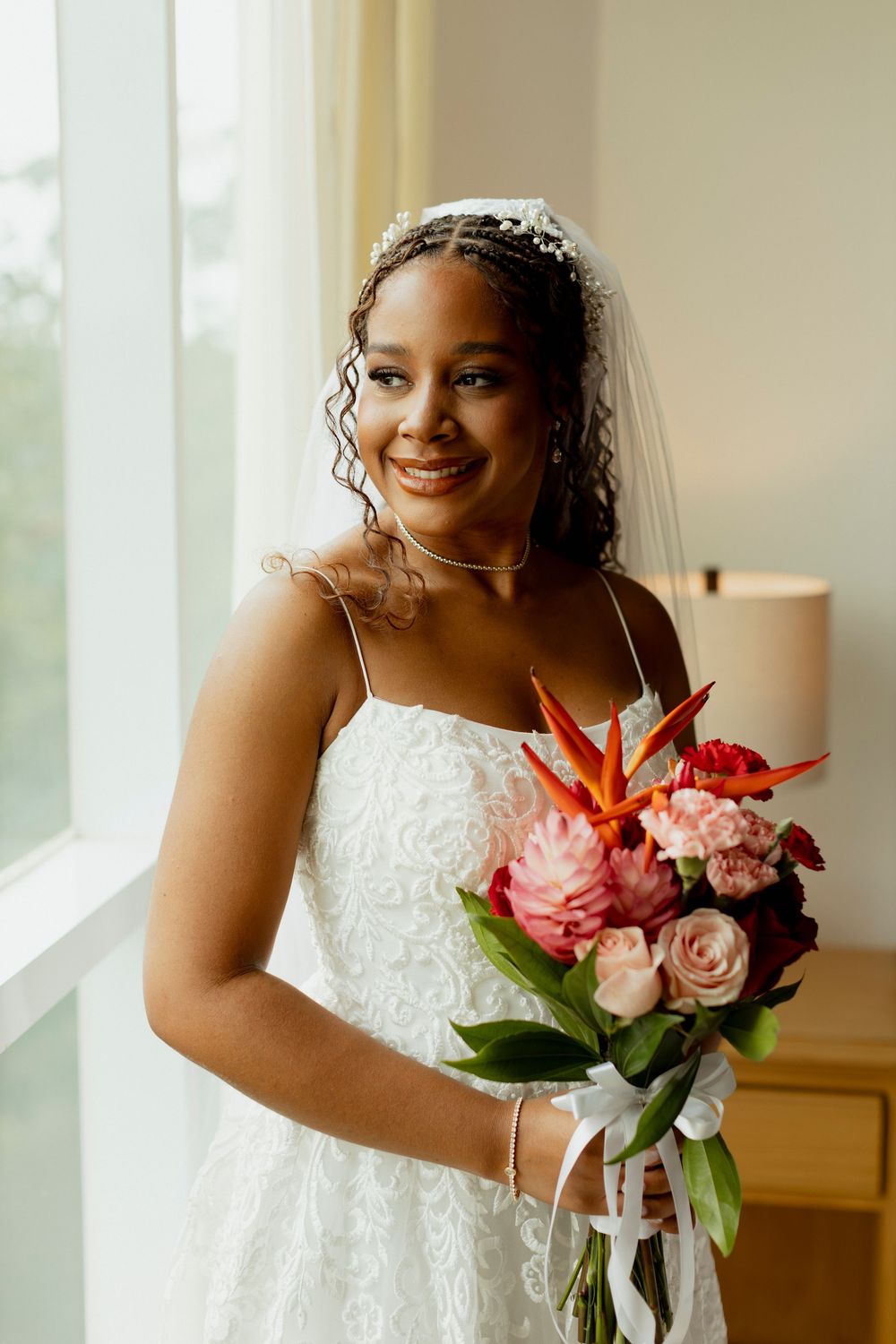Smiling bride in white lace gown and veil holds vibrant tropical bouquet with roses and bird of paradise flowers.