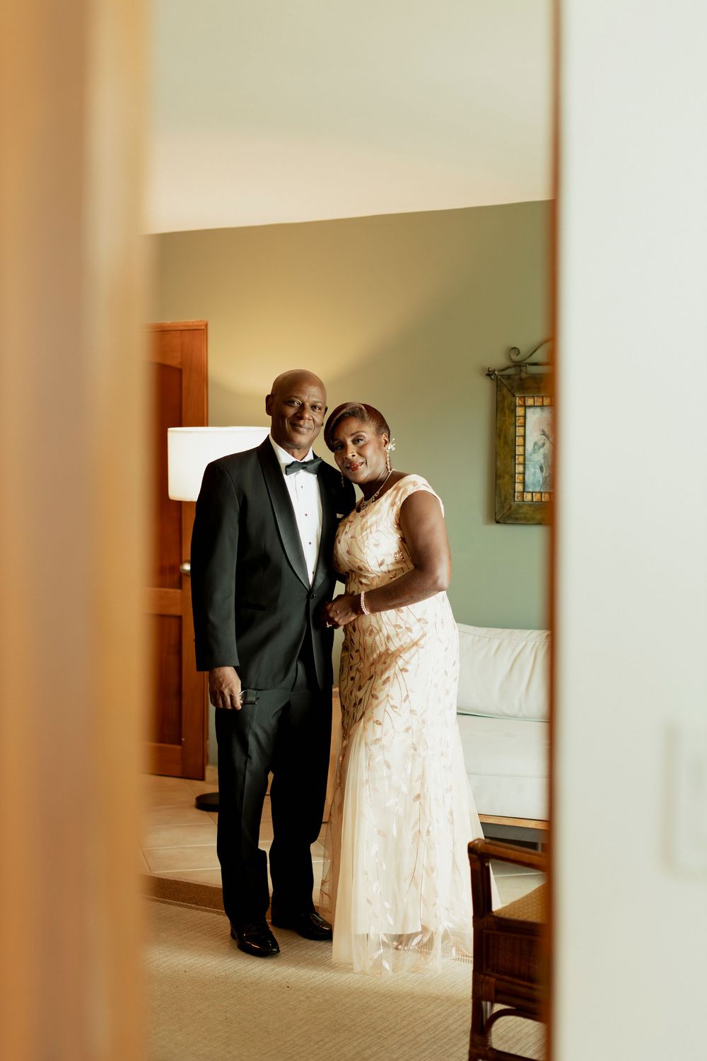 Elegant couple in wedding attire posing in a hotel room, man in tuxedo and woman in lace gown.
