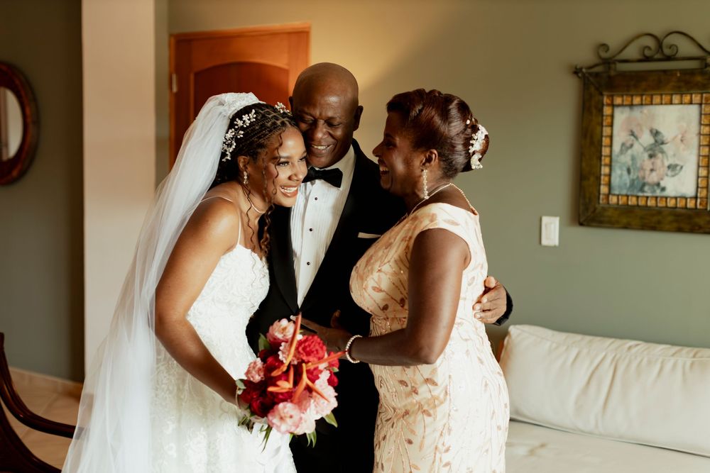 Bride in white lace gown and veil holds red bouquet while sharing tender moment with man in tuxedo and woman in gold dress.