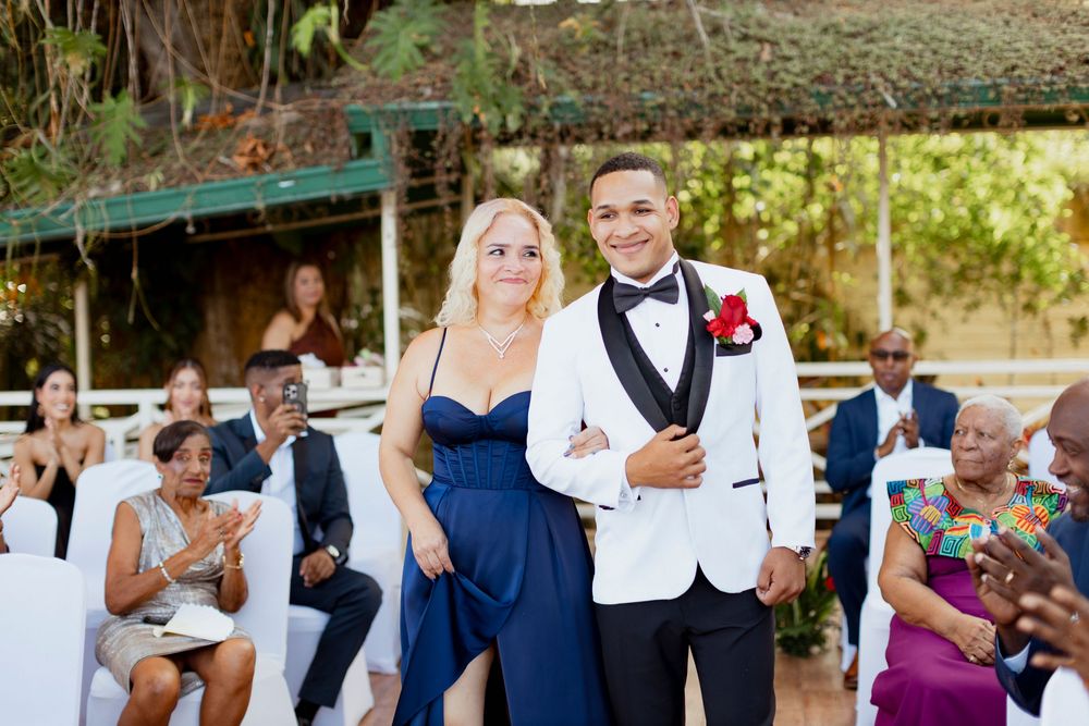Elegant couple walking down the aisle at an outdoor wedding ceremony, man in white tuxedo and woman in navy blue gown.