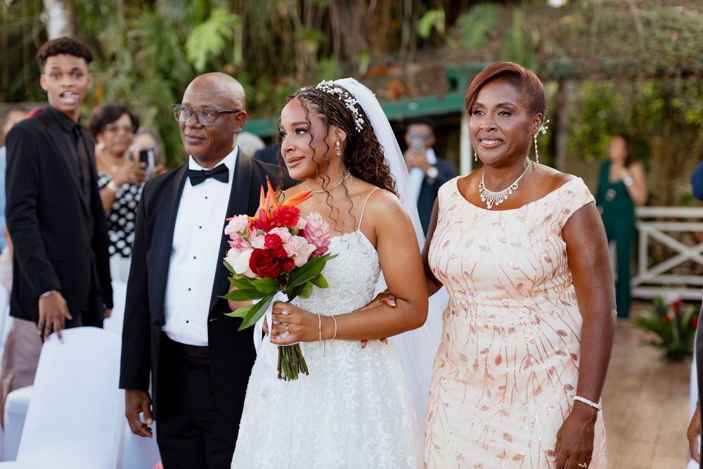 Bride in white gown holding colorful bouquet walks down aisle flanked by parents at outdoor wedding ceremony.