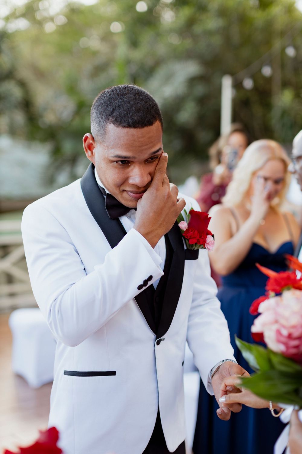 Emotional groom in white tuxedo wiping tears during outdoor wedding ceremony with guests behind.