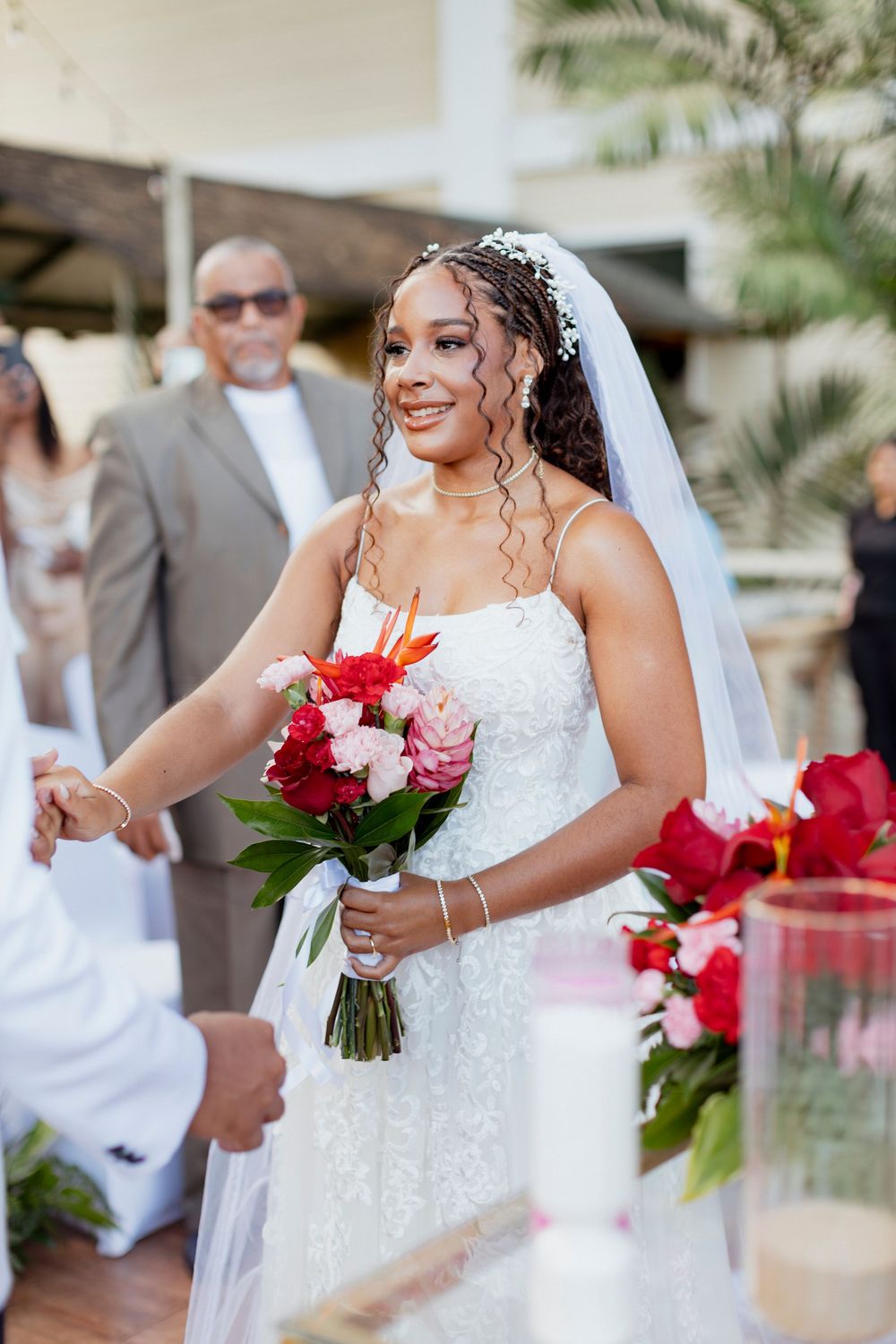Smiling bride in white lace gown and veil walks down aisle holding red and pink bouquet at outdoor wedding ceremony.
