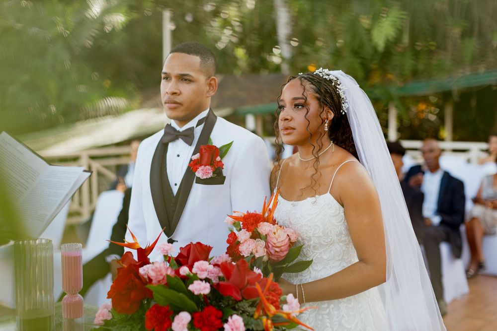 A couple stands at the altar during an outdoor wedding ceremony, the bride in white holding a red and pink bouquet.