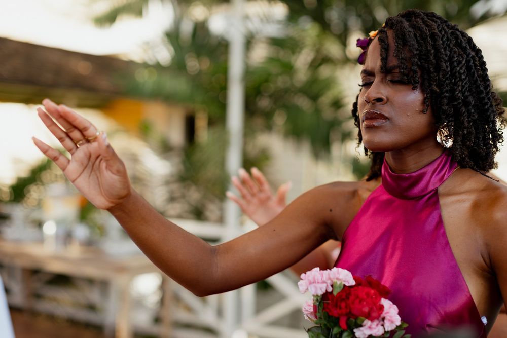 A woman in a pink halter dress raises her hands while holding a bouquet of pink and white flowers outdoors.