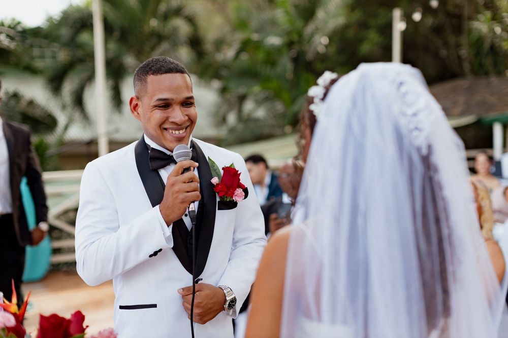 Groom in white tuxedo jacket speaks into microphone during outdoor wedding ceremony facing his bride in white veil.