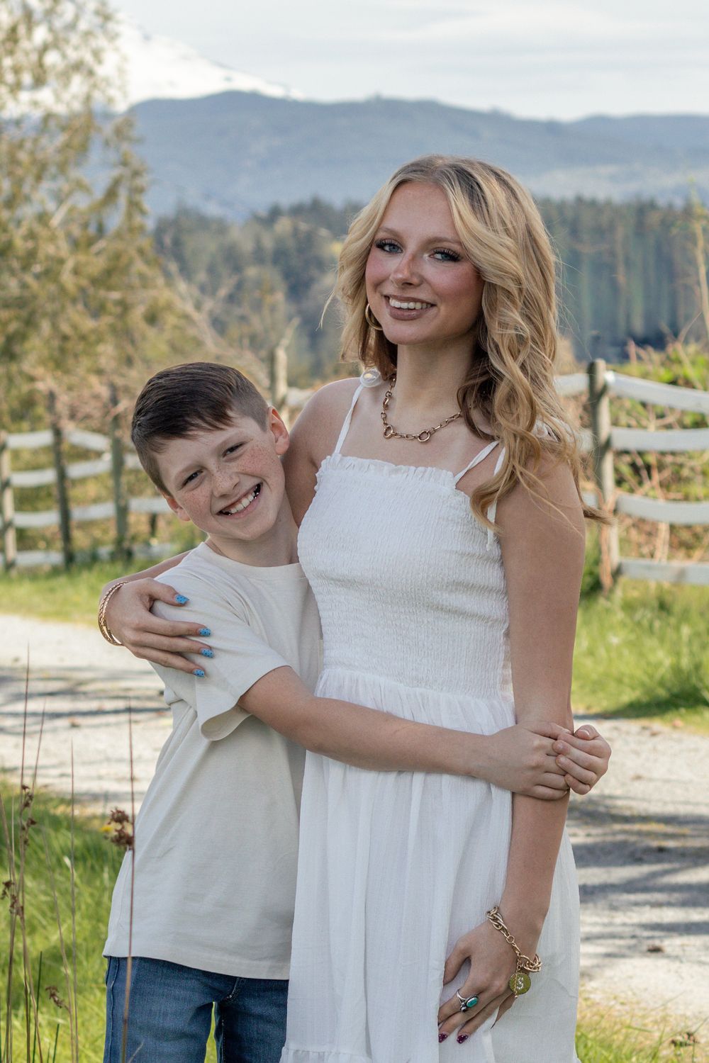 Smiling teenage girl in white dress hugging a laughing young boy outdoors near a wooden fence in eatonville washington taken by photographer