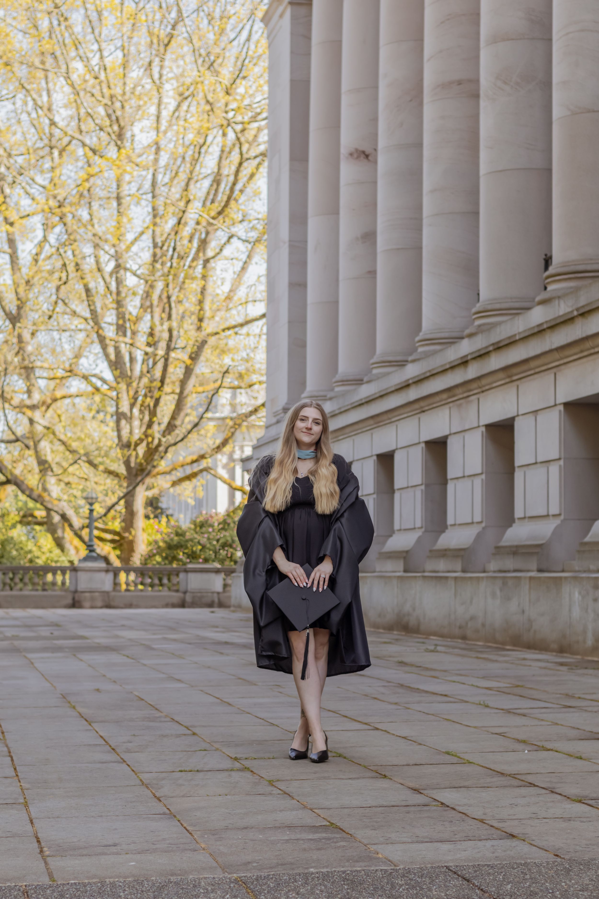 college Graduate in black cap and gown walking beside large stone columns on Washington capitol campus taken by olympia photographer