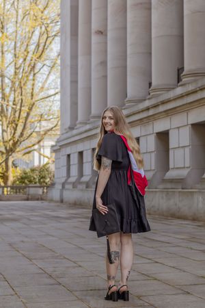Smiling graduate in black dress with red masters degree hood looking back over her shoulder outside Washington capitol building.