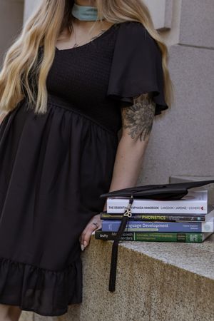 Blonde woman in black dress holding graduation cap and speech-language pathology textbooks on stone steps at Washington capitol building
