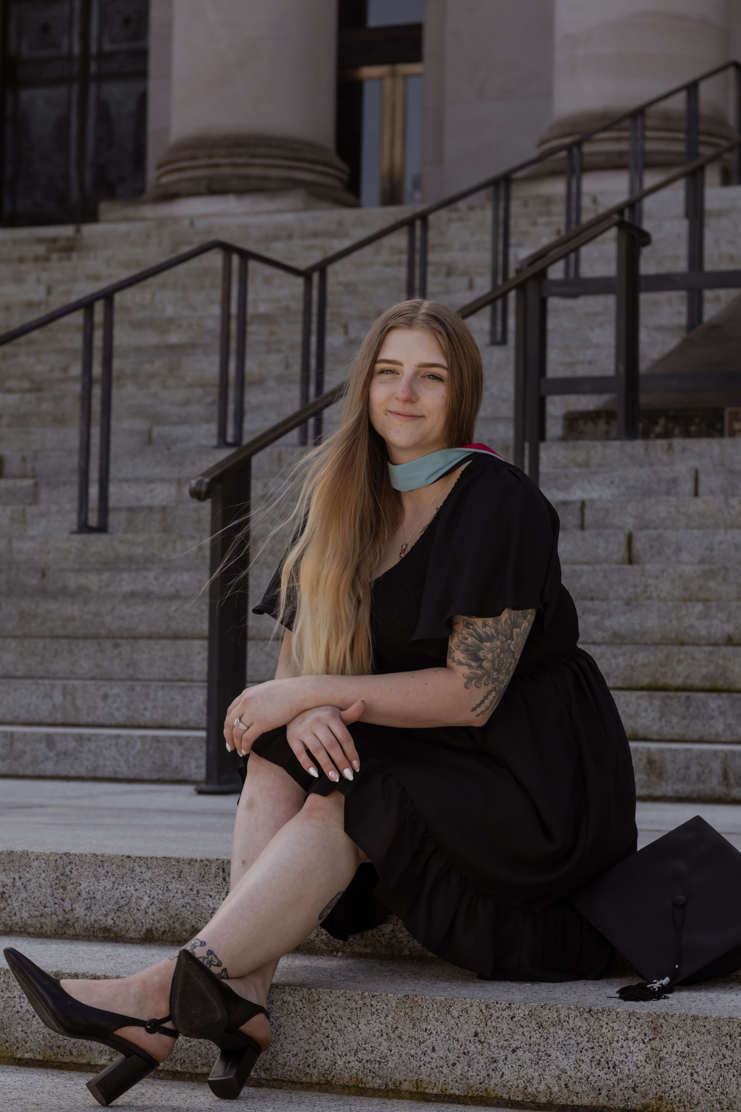 Smiling tattooed blonde woman in black dress sitting on Washington capitol steps with graduation cap beside her taken by Roy photographer