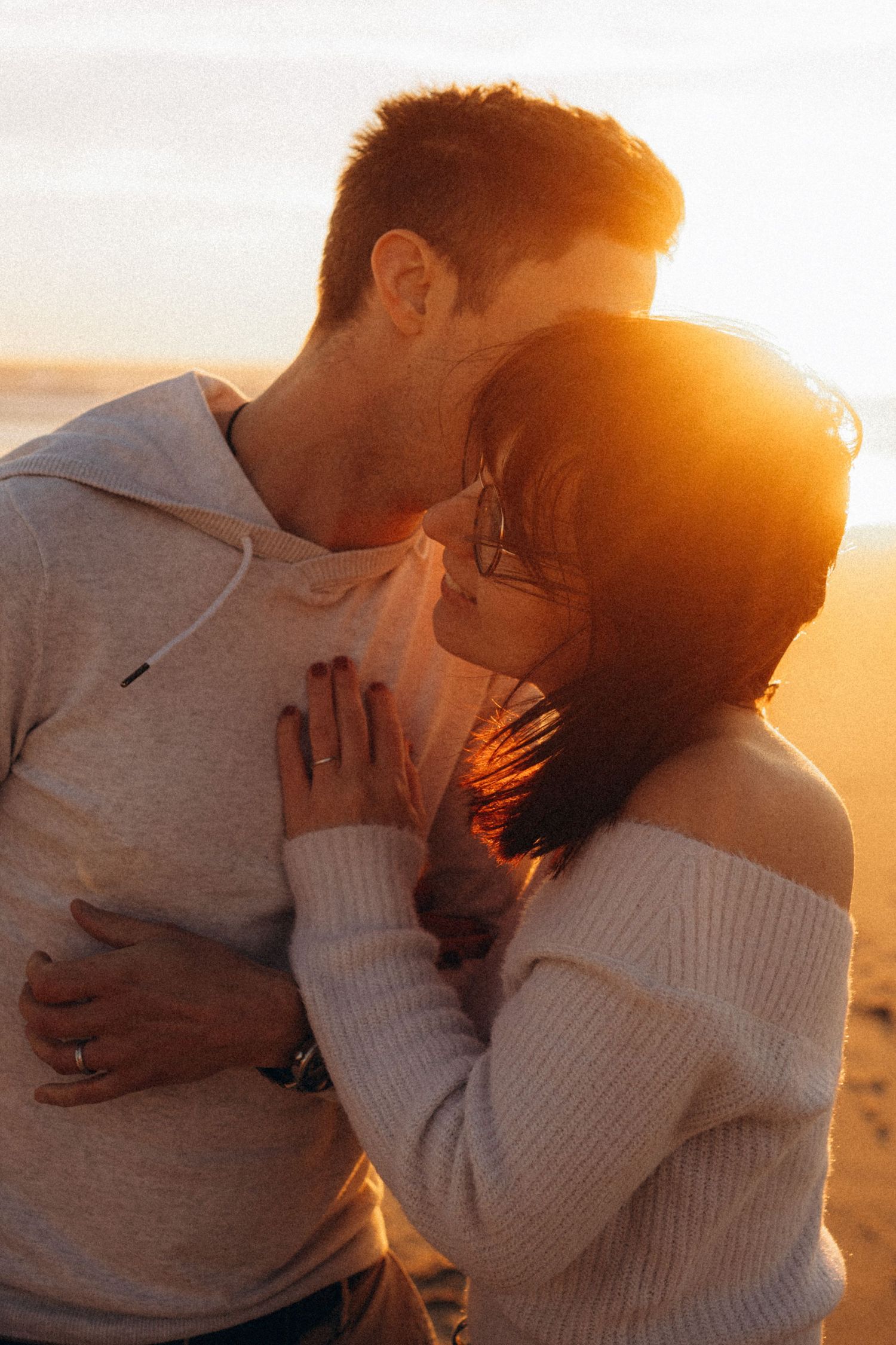 Couple sharing a romantic moment at golden sunset on the beach, woman in off-shoulder sweater embracing man in hoodie.