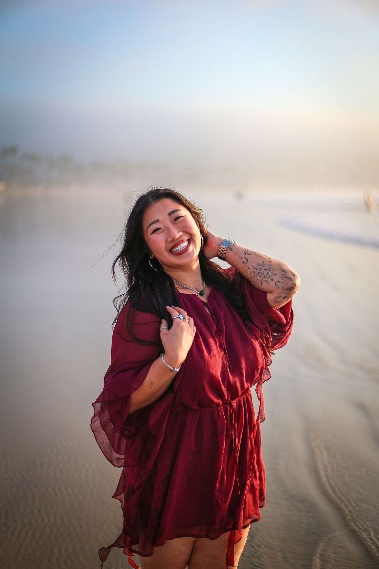 Joyful Asian woman in burgundy dress laughing on foggy beach during soft golden sunset light.