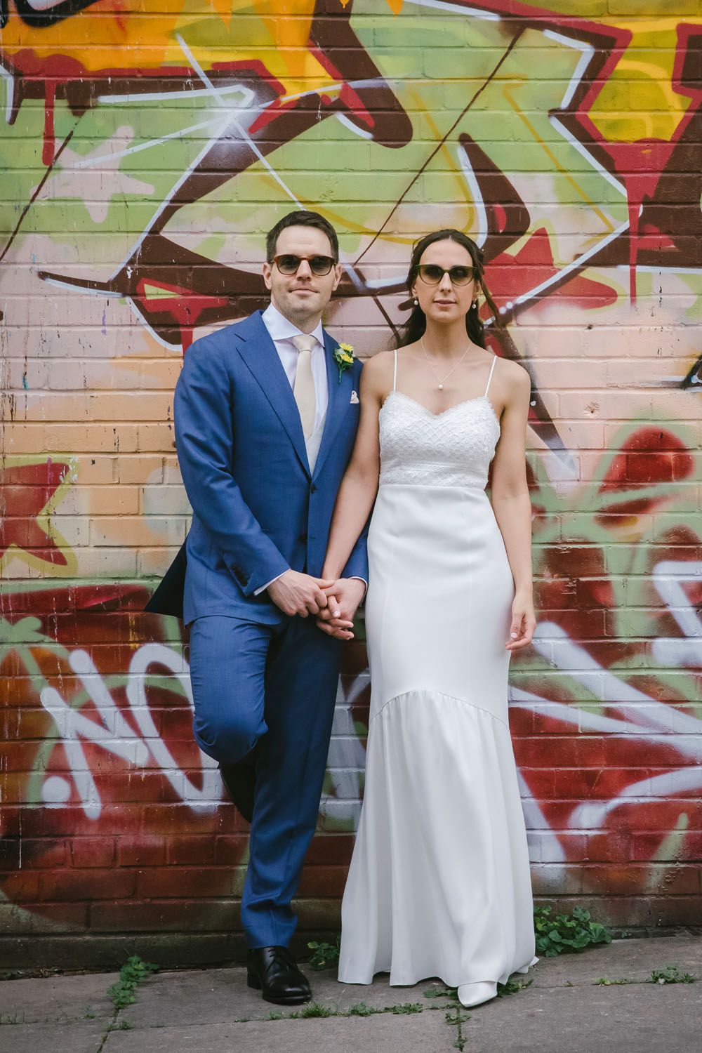 A bride and groom in sunglasses pose holding hands in front of a colorful graffiti wall, wearing white dress and blue suit.
