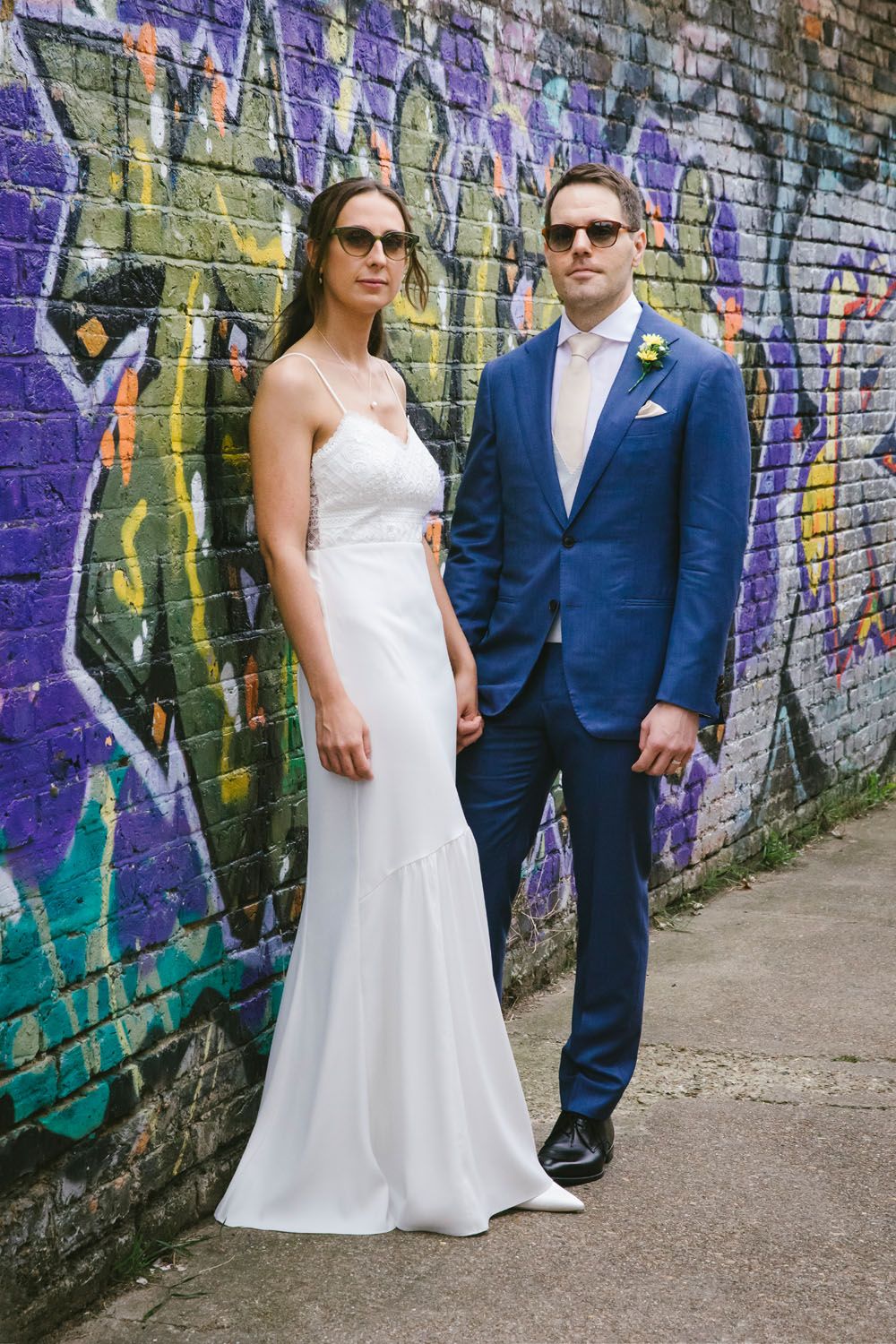 Bride in white gown and groom in blue suit wearing sunglasses pose in front of colorful graffiti wall.
