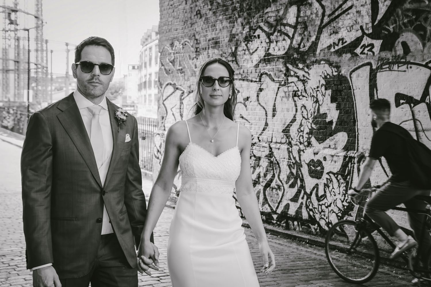 Black and white photo of a bride and groom holding hands in front of a graffiti wall, with a cyclist passing by.